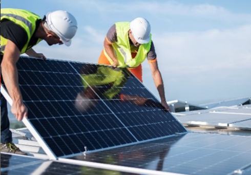 Two workers are installing solar panels on a roof, wearing safety vests and helmets as they lift a panel.