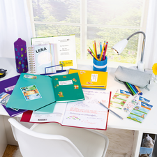 Ein Schreibtisch mit bunten Ordnern, Schreibmaterialien, einer Lampe und einer Blume. Im Hintergrund sind Fenster und ein farbenfrohes Aufbewahrungselement sichtbar.