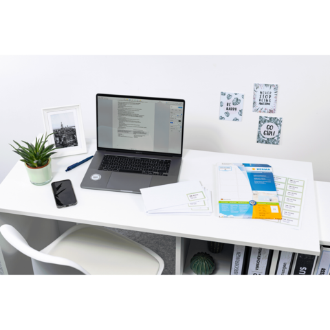 Schreibtisch mit Laptop, Smartphone und Pflanzen. Dokumente und Notizen liegen auf dem Tisch, während Bilder an der Wand hängen.