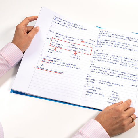 A person is holding an open notebook filled with handwritten notes and markings in blue and red ink.