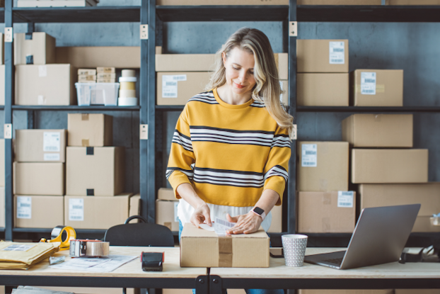 A woman stands at a table, surrounded by boxes and a laptop, in a room filled with shelves of packages.