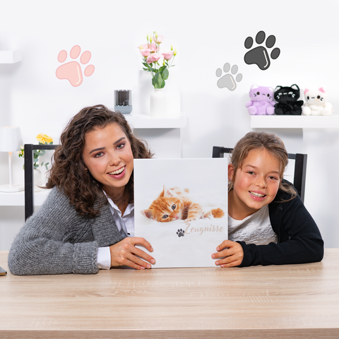 A woman and a girl are holding a book with a kitten cover while sitting at a table. Decorative elements are visible in the background.