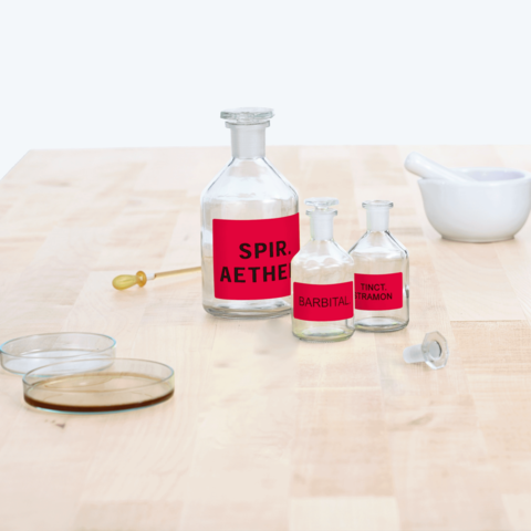 Flaschen mit roten Etiketten, beschriftet mit "Spir. Aether", "Barbital" und "Tinct. Tramon", sowie ein Mörser und Petrischalen auf einem Tisch.
