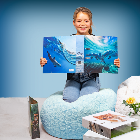 A girl is sitting on a beanbag chair, holding a book with pictures of dolphins. Next to her, there are more books on a table.