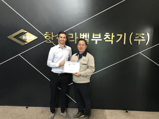 Two men are holding a certificate in front of a wall with the inscription 'HSLM' and Korean characters.