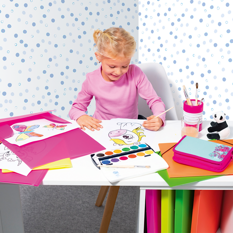A little girl sits at a table, painting with watercolors, surrounded by colorful paper and drawings.