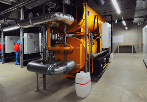 An orange heating boiler with pipes and tanks in a technical room, surrounded by white appliances and a work surface.
