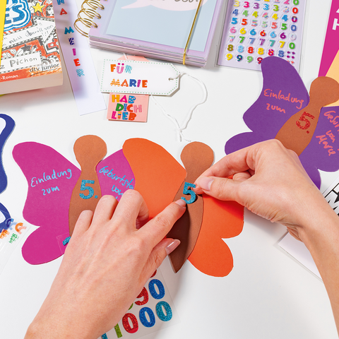 Colorful invitations in the shape of butterflies are designed with numbers and text. Hands add the finishing touches.
