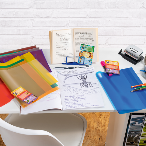 Colorful plastic folders, a stack of notes, and a binder on a table. In the background, an open book with handwritten notes.