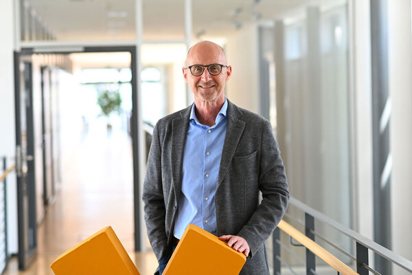 A man with glasses stands in a modern office hallway, holding two orange cushions. He is wearing a gray blazer and a blue shirt.