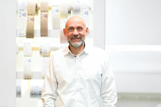A man with a beard and a bald head is wearing a white shirt and standing in front of a wall with various color samples.