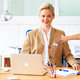 A woman with blonde hair is sitting at a desk, wearing a blazer and smiling. In front of her is a laptop and some writing materials.