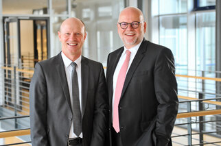Two men in suits are smiling in a modern office building. The man on the left is wearing a tie, while the one on the right has a lighter tie.