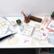 Schreibtisch mit Planer, Schreibutensilien, einer Pflanze und verschiedenen Dekorationselementen. Farbiges Papier und Aufkleber sind verteilt.