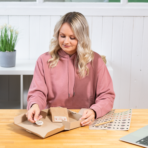 A person with blonde, curly hair is sitting at a table, working with a cardboard box and colorful stickers.
