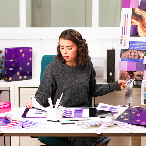 A person is sitting at a table, surrounded by color swatches and design materials, as they work on a creative project.