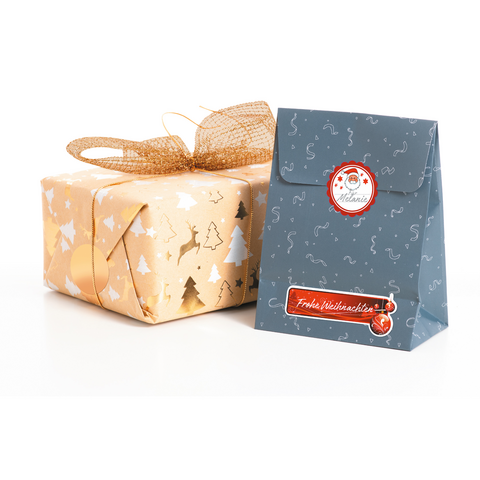 Geschenkpaket mit goldener Schleife und weihnachtlichem Motiv, daneben eine dunkelblaue Tüte mit dem Aufdruck „Frohe Weihnachten“.