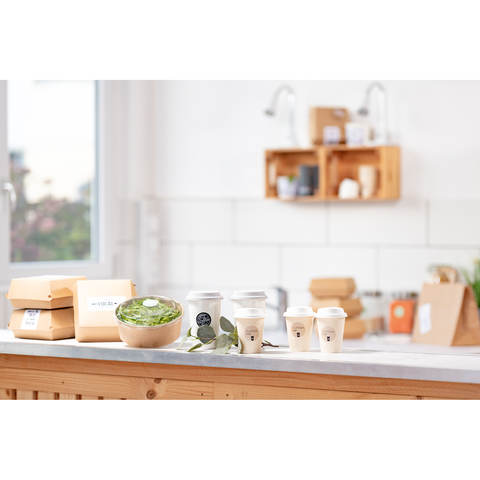 Verschiedene Takeaway-Behälter, darunter eine Schüssel mit Salat und mehrere Kaffeebecher, auf einer Holzoberfläche in einer hellen Küche.
