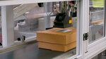 A conveyor belt transports two brown boxes while a machine processes them. A label is visible on one of the boxes.