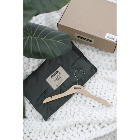 Ein schwarzes Paket mit einem Aufkleber liegt auf einer weißen Decke, daneben ein Holzkleiderbügel und eine Pflanze im Hintergrund.
