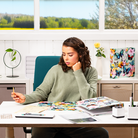 A young woman sits at a desk, surrounded by colorful materials and notes, as she thoughtfully holds a pen.