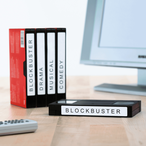 Eine Sammlung von VHS-Kassetten mit den Beschriftungen "BLOCKBUSTER", "DRAMA", "MUSICAL" und "COMEDY" neben einer Fernbedienung und einem Computer.