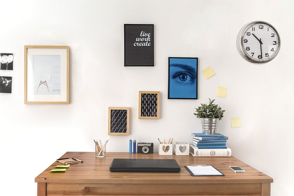 A desk with a laptop, decorative pictures on the wall, a plant, and a floor lamp. A wall clock shows the time.