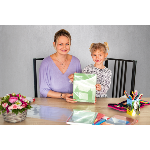 Eine Frau und ein Mädchen sitzen an einem Tisch, halten ein grünes Heft und lächeln. Auf dem Tisch liegen Schreibwaren und ein Blumenstrauß.