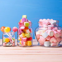 Colorful candies in jars, including lollipops, gummy bears, and heart-shaped marshmallows, set against a light wooden background.