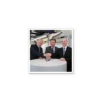 Three men in suits stand around a table, placing their hands on an object in the center.