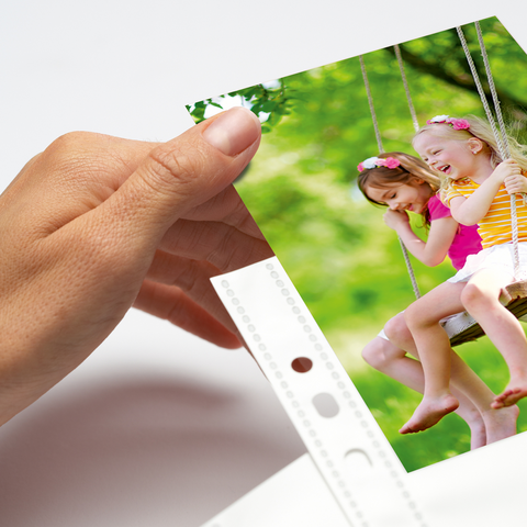 A hand holds a photo of two girls playing on a swing outdoors, surrounded by trees and green grass.