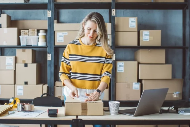 A woman in a striped sweater stands at a table, surrounded by boxes and a laptop in a modern office.