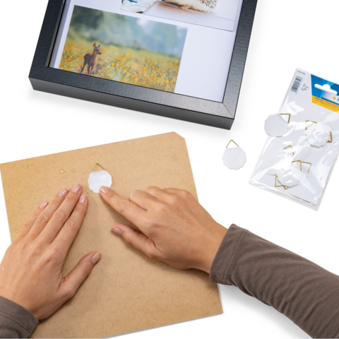 Eine Hand platziert eine weiße Form auf einem braunen Untergrund, während eine Bilderrahmen und eine Packung mit Clips im Hintergrund sichtbar sind.