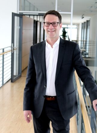 A man in a suit stands in a modern office hallway, smiling as he holds onto a railing.