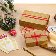 Geschenkpäckchen in goldener Verpackung mit roten Bändern, umgeben von Geschenkpapier, Aufklebern, einer Tannenzapfen und Kerzenhalter.