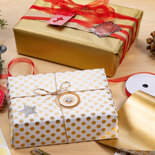 Geschenkpäckchen in gold und weiß mit Punkten, umgeben von Dekorationen wie Kerzen, einer Tannenzapfen und Geschenkband.