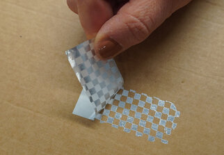 A hand peels off a piece of transparent, checkered tape from a cardboard surface.
