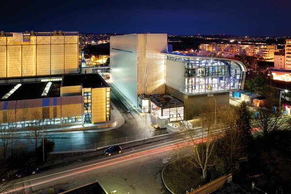 Modern architecture of a building at night, surrounded by city lights and a street filled with vehicle headlights.