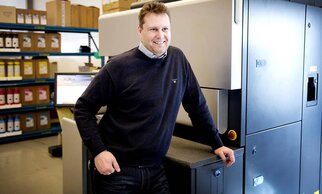 A man stands smiling next to a printing press in a modern workspace, surrounded by shelves filled with printing materials.
