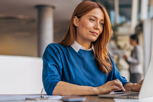 A woman with long red hair is sitting at a table, working intently on a laptop in a modern room.