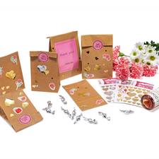 Geschenktüten mit Aufklebern, eine Flasche Sekt und Blumenarrangement. Dekorative Elemente und Dankeskarten sind ebenfalls sichtbar.