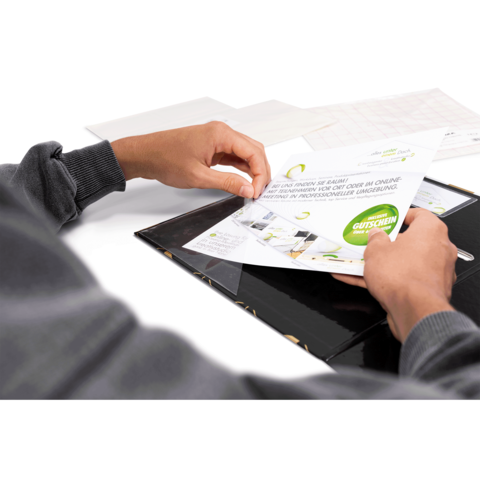 Eine Hand hält einen Gutschein in einem Ordner, während eine andere Hand das Dokument leicht anhebt. Der Hintergrund ist neutral und unscharf.