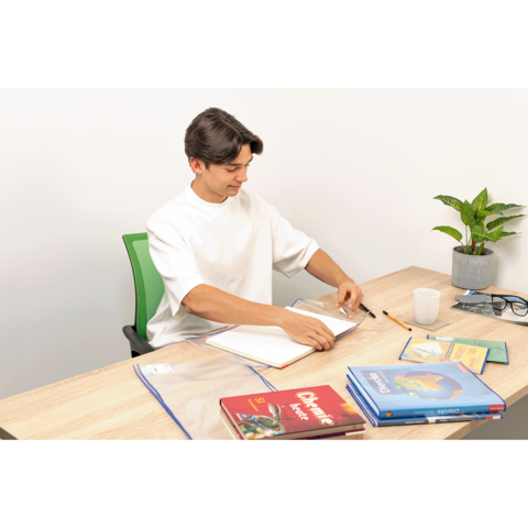 Ein junger Mann sitzt an einem Tisch, blättert in einem Notizbuch und hat Bücher sowie Schreibutensilien vor sich. Eine Pflanze steht daneben.