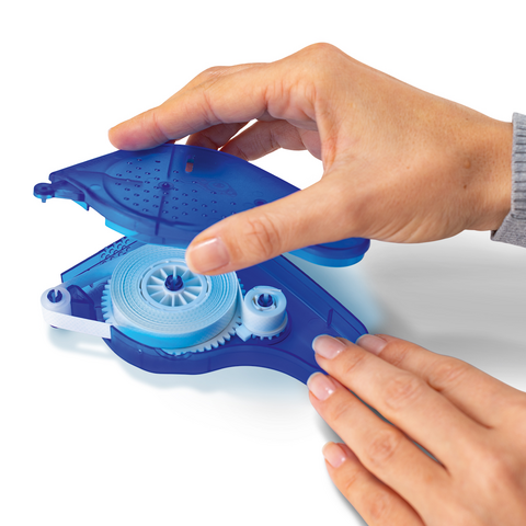 A hand holds a blue correction tape dispenser while the other hand operates the mechanism. The dispenser is partially open to reveal its inner workings.