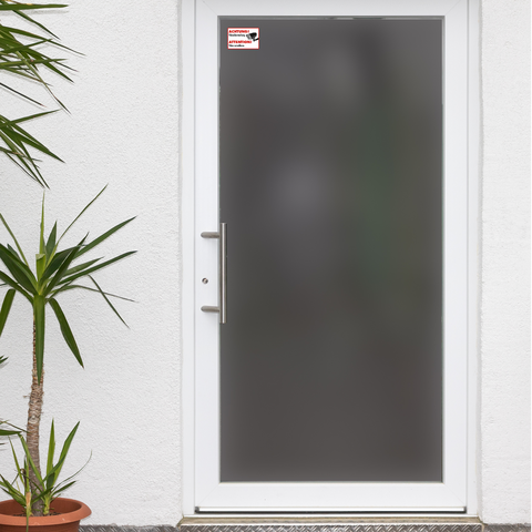 Weiße Eingangstür mit grauem Glas und modernem Türgriff, umgeben von einer Topfpflanze. Ein Warnschild ist oben rechts angebracht.