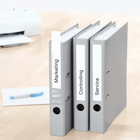 Drei graue Ordner mit den Beschriftungen "Marketing", "Controlling" und "Service" stehen auf einem Tisch, daneben ein blauer Kugelschreiber.