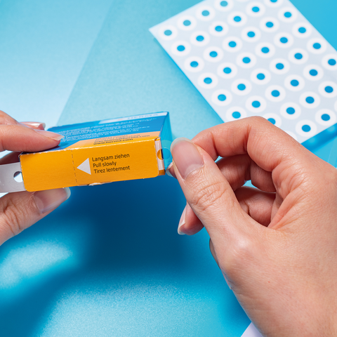 Eine Person hält eine Verpackung mit einem gelben Etikett in der Hand, während sich im Hintergrund ein Blatt mit Löchern und Diagramme befinden.