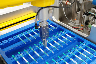 An automated robotic arm picks up a syringe from a blue tray containing several syringes that are neatly arranged.