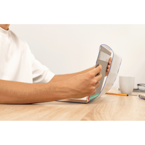 Eine Hand hält ein Smartphone, während die andere ein aufgeschlagenes Notizbuch auf einem Tisch stützt. Im Hintergrund steht eine Tasse.