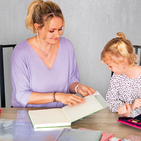Eine Frau und ein Mädchen sitzen an einem Tisch und betrachten gemeinsam ein Buch. Auf dem Tisch liegen verschiedene Schreibmaterialien und Blumen.